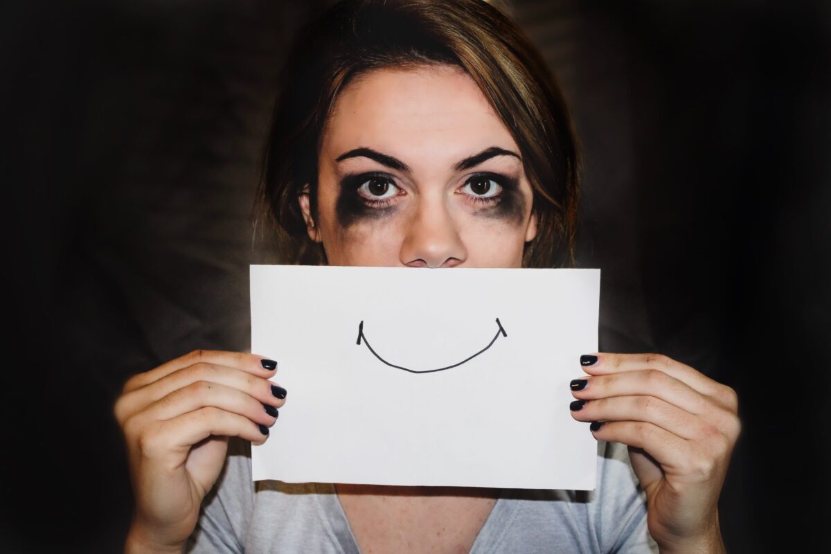 A person holding a sheet of paper with a smile drawn on it in front of their mouth, with smudged mascara under their eyes
