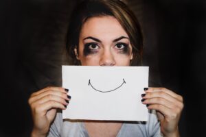 A person holding a sheet of paper with a smile drawn on it in front of their mouth, with smudged mascara under their eyes
