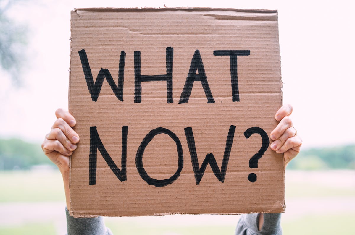 Cardboard sign reading 'What Now?' held outdoors, conveying uncertainty or protest.