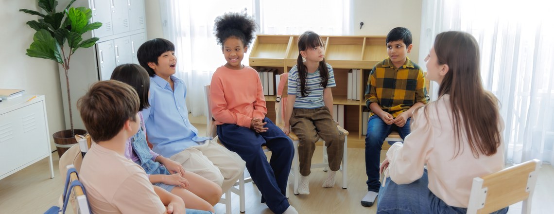 A group of school-age children and a teacher sitting in a circle of chairs in a bright classroom