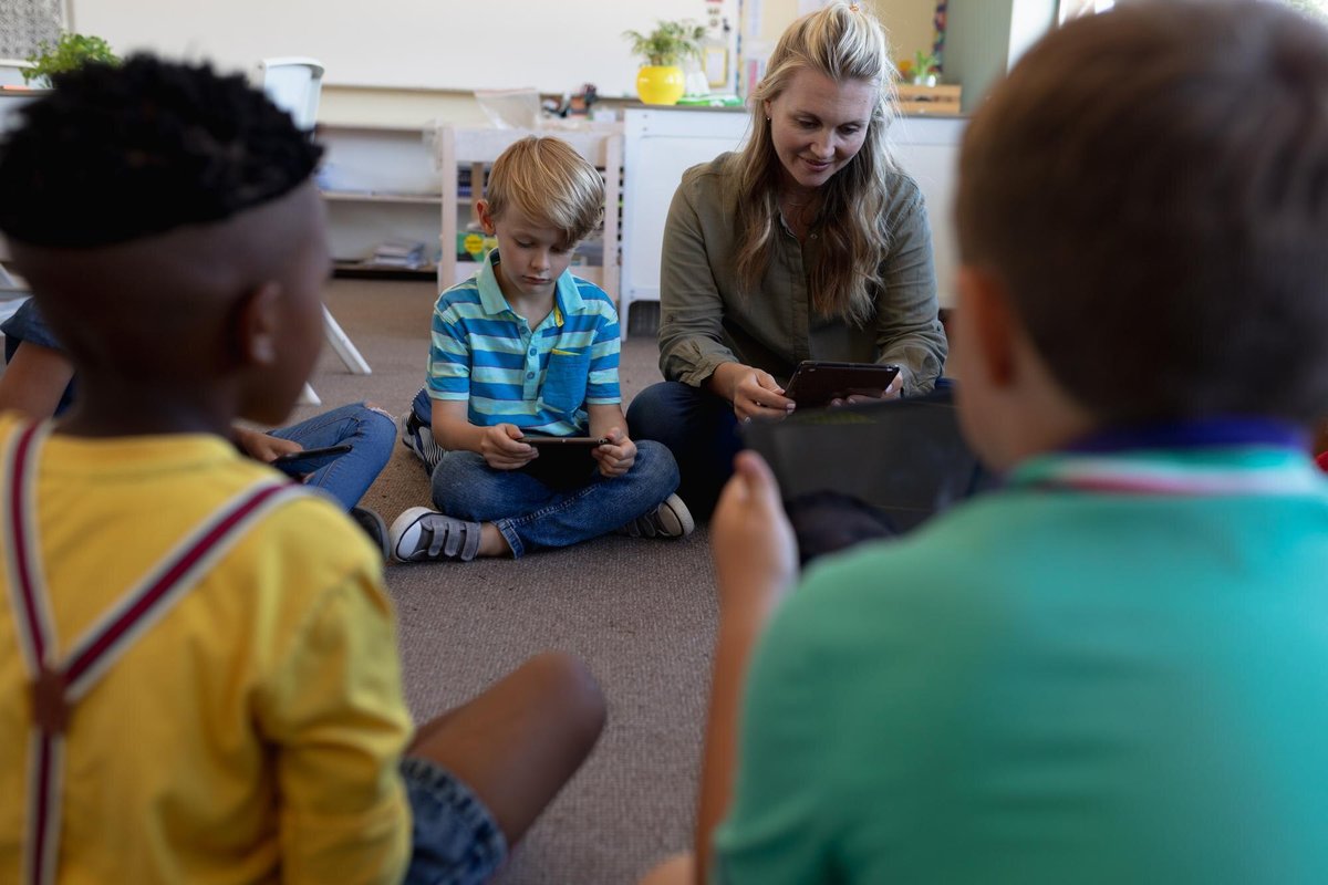 A teacher and children sitting on the floor of a classroom, looking at tablets together