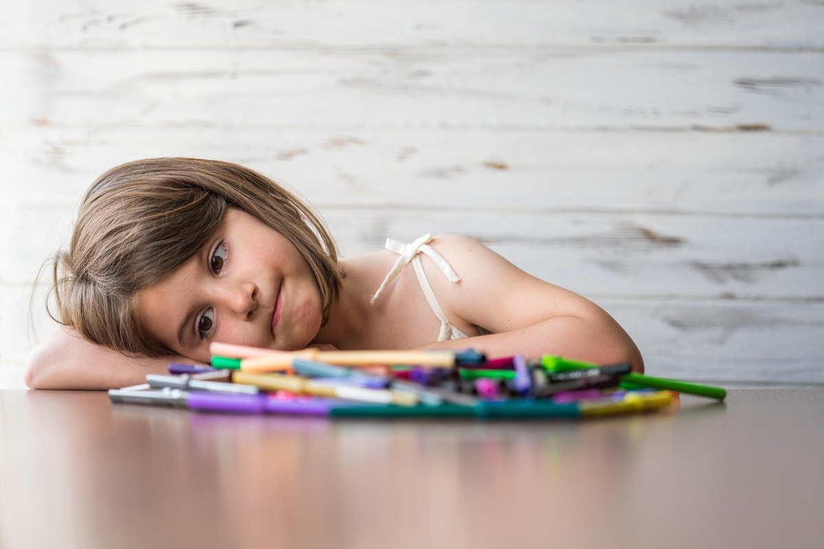 A young child resting her head on a table beside a pile of felt-tip pens