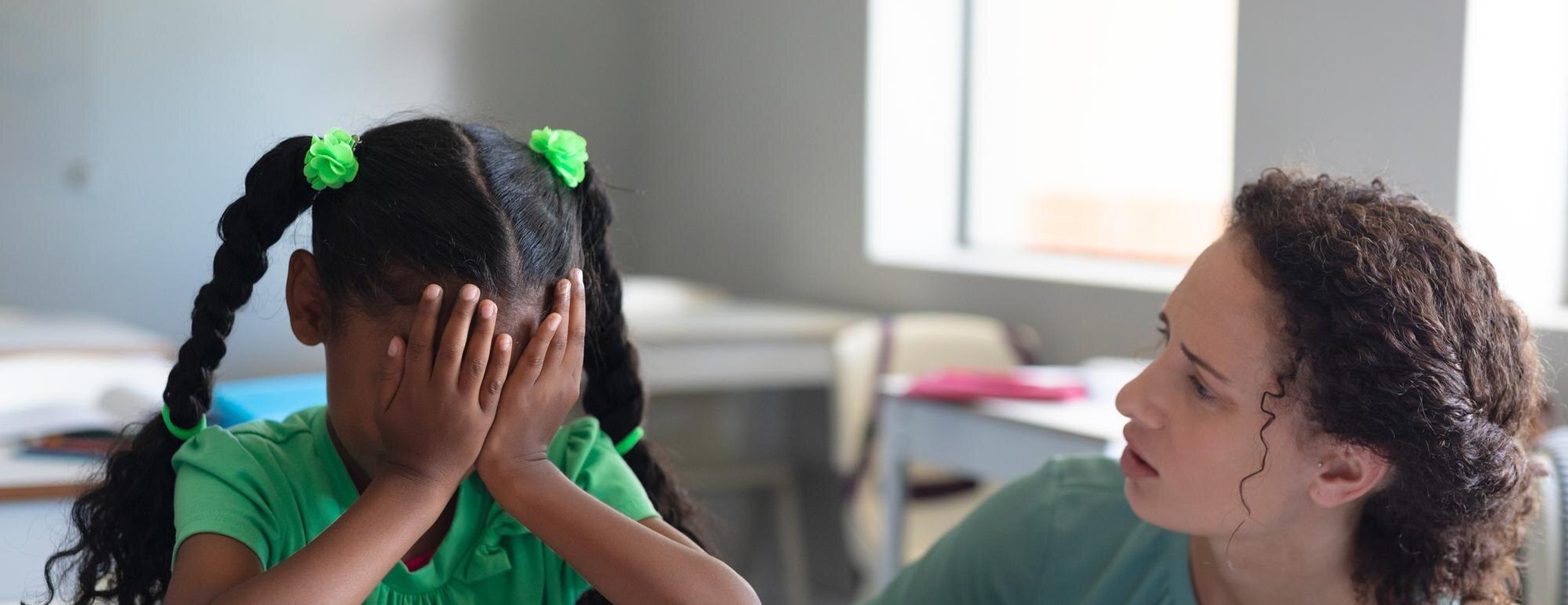 A child sitting at a school desk with her hands over her face while a teacher speaks with her
