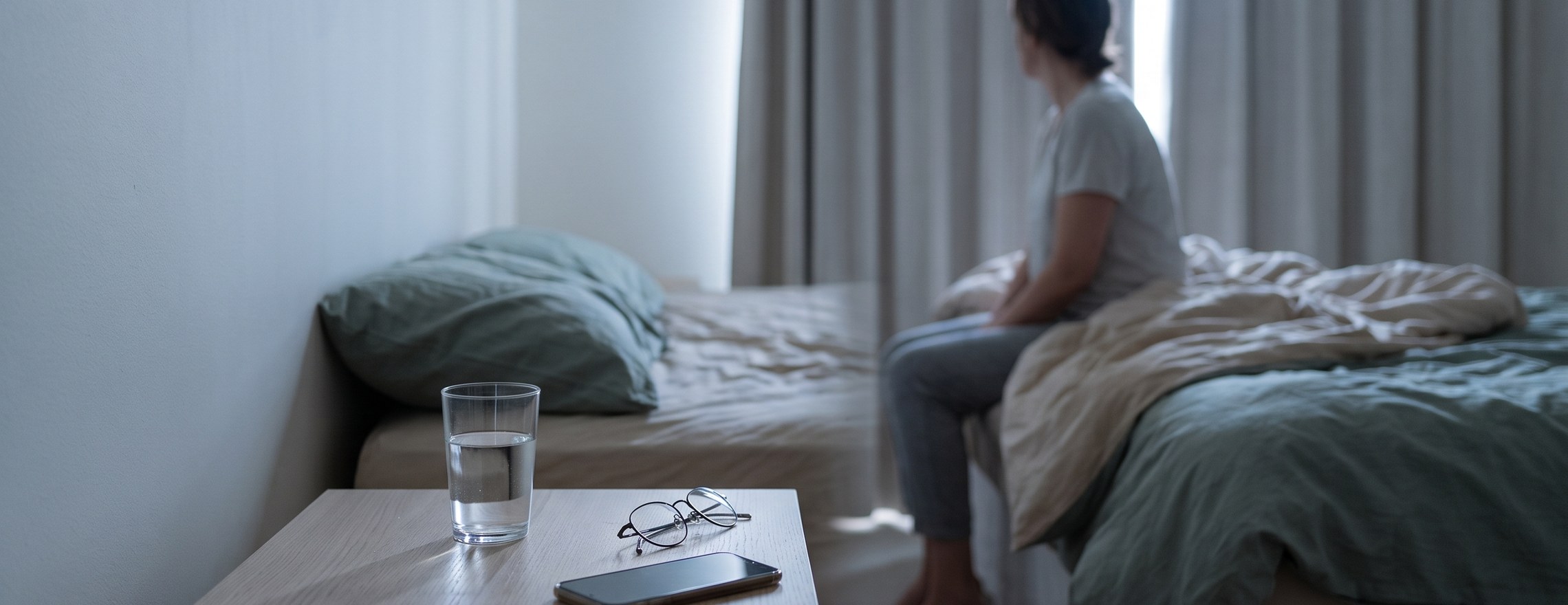 A bedside table with a glass of water and glasses in the foreground, and a person sitting on the edge of an unmade bed by the window