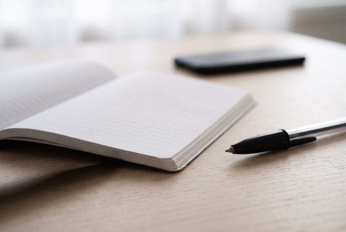 A phone lying face down on a table beside an open notebook and pen