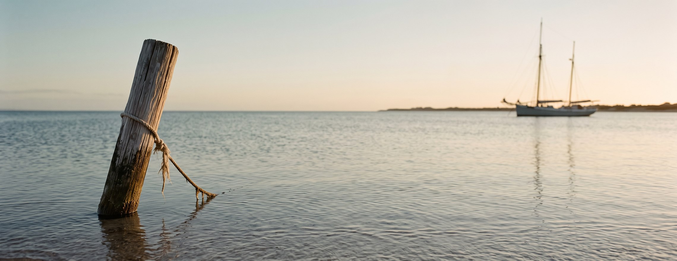 A weathered mooring post in shallow water at sunset, with a two-masted sailing boat at anchor further out, out of focus