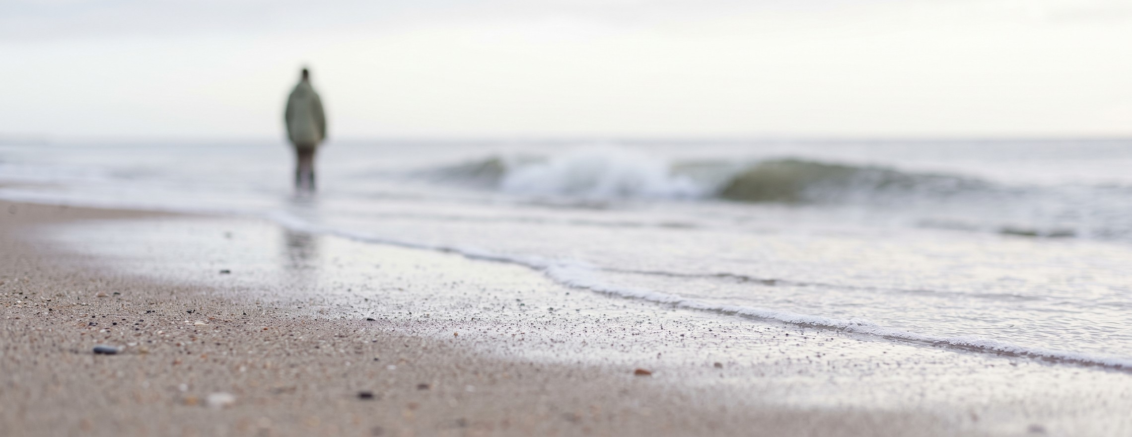 The edge of a spent wave on wet sand, with a breaking wave and a lone figure standing at the water’s edge behind