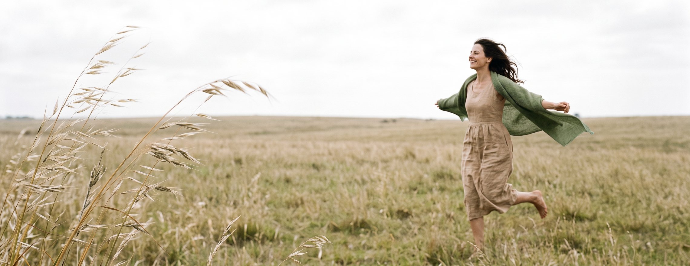 A person moving through long dry grass in an open field, a light shawl lifting in the wind behind them
