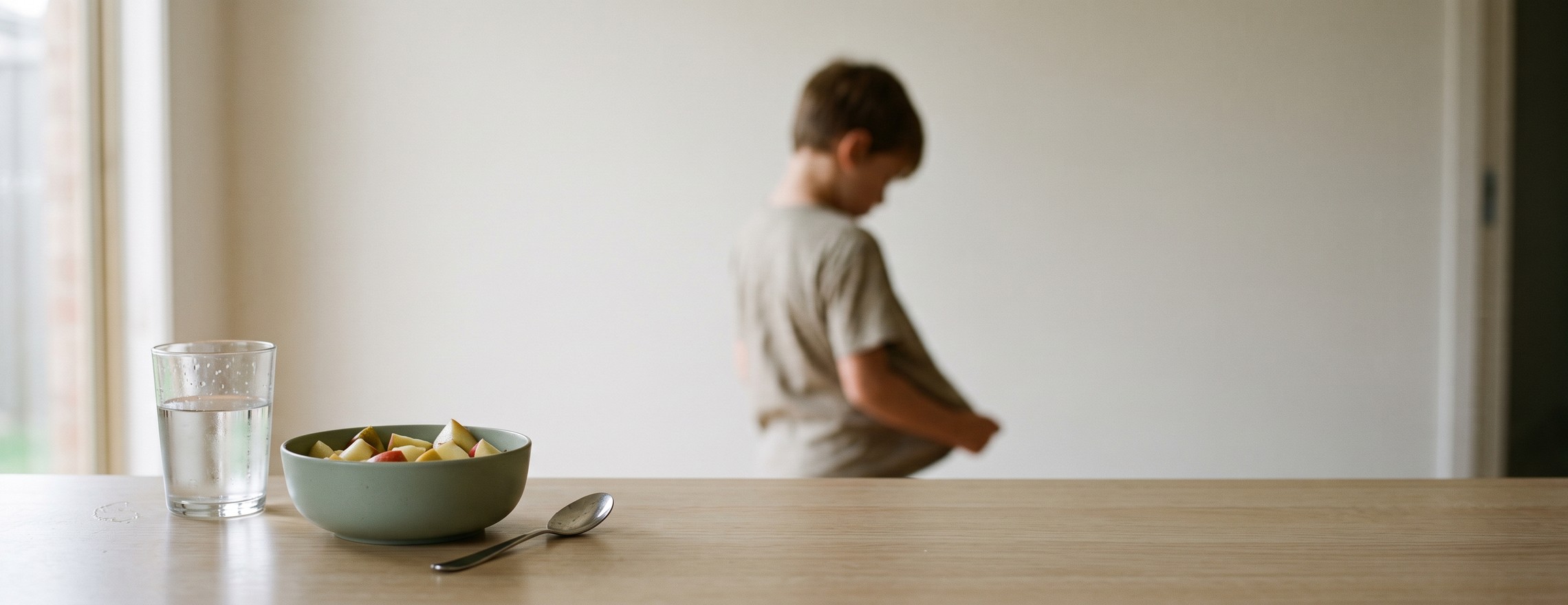 A bowl of cut fruit and a glass of water untouched on a kitchen bench, with a child standing further back turned away, out of focus