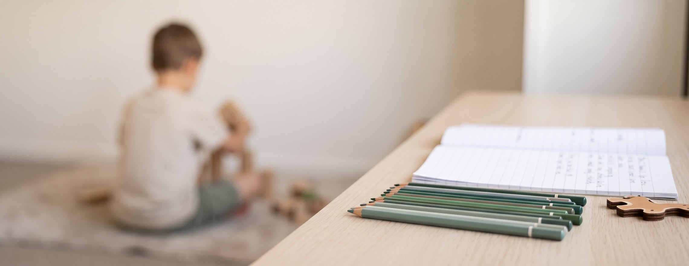 A desk with coloured pencils and an open exercise book in the foreground, and a child sitting on the floor building with wooden blocks behind