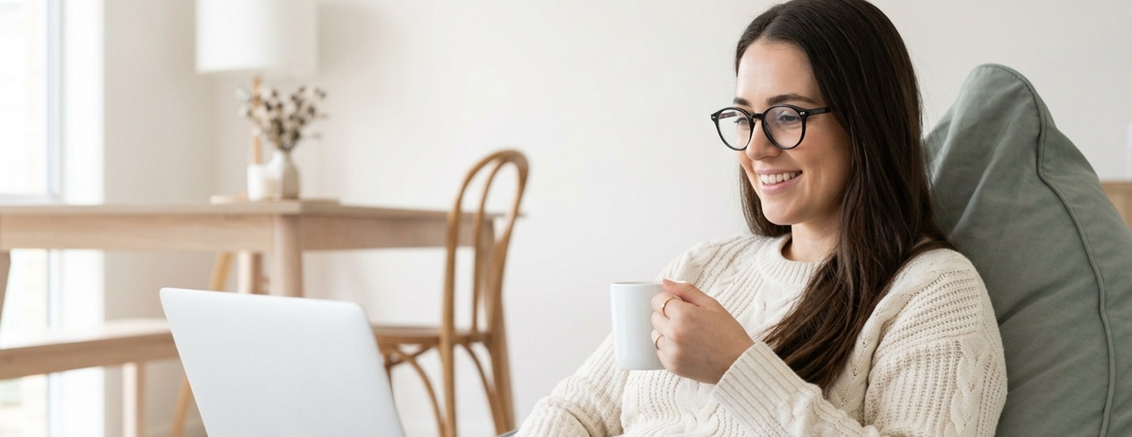 A woman sitting comfortably in a beanbag at home with a laptop and a mug, part way through a telehealth session