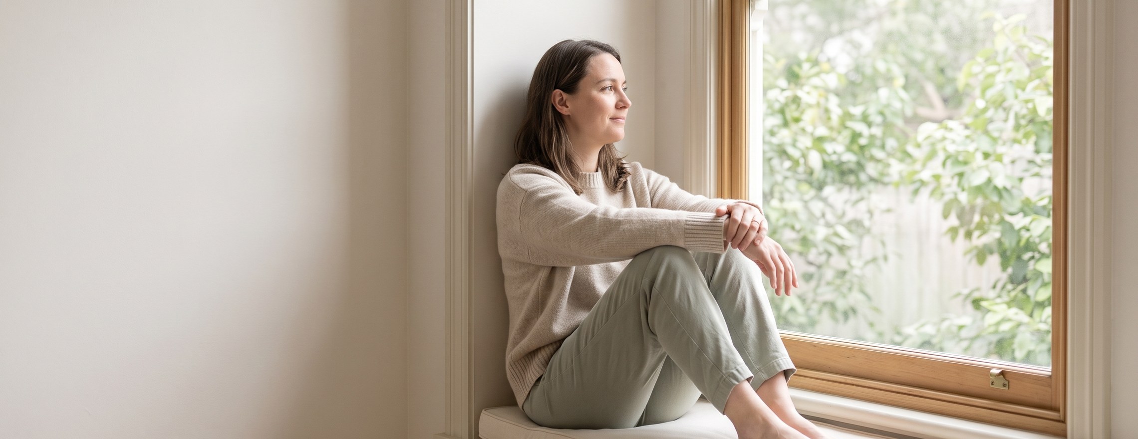 A woman sitting on a window seat at home, looking out at a green garden with a calm, quiet expression