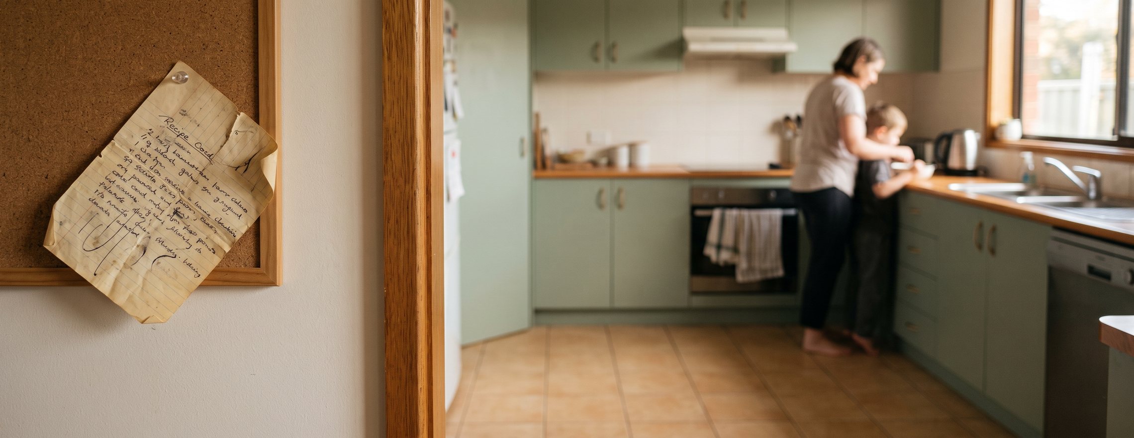 A handwritten recipe card pinned to a corkboard beside a doorway, with an adult and a child cooking together at the kitchen bench, out of focus