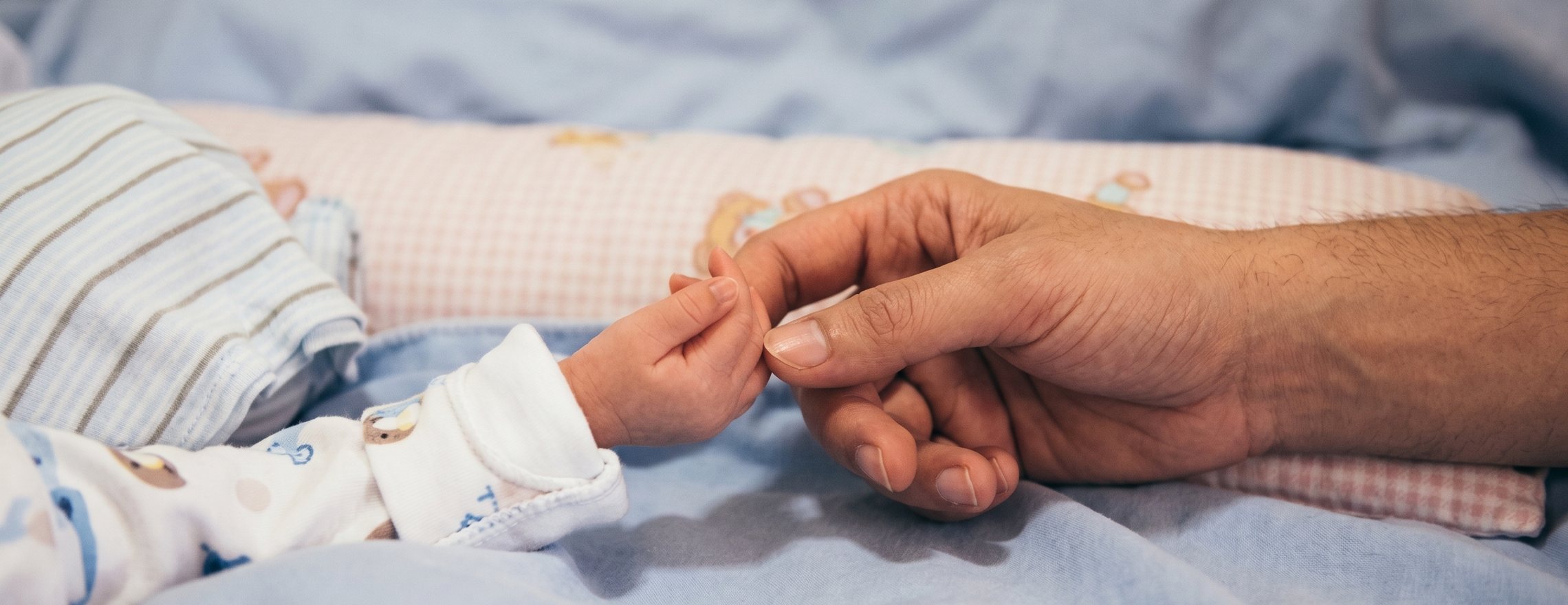 A newborn baby’s hand curled around an adult’s finger, resting on a cot sheet in soft daylight