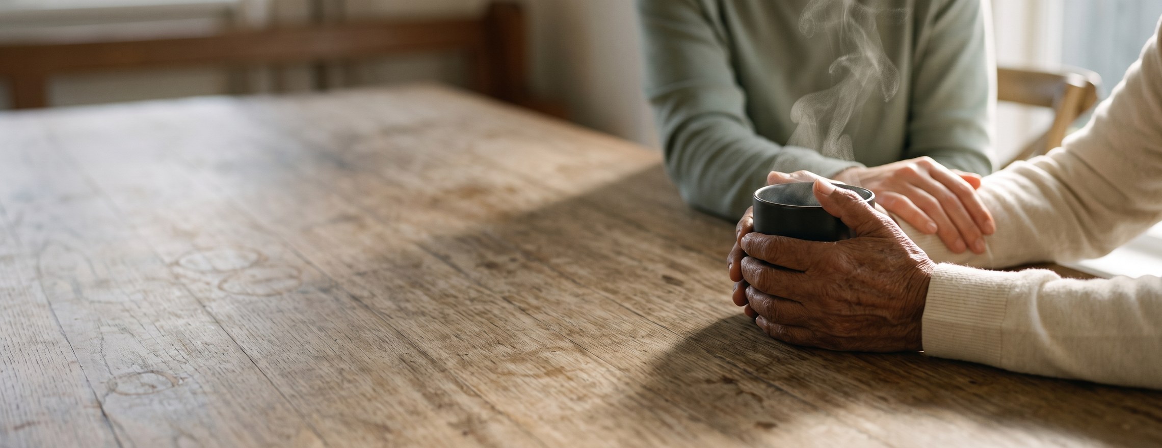 Two pairs of hands on a worn timber kitchen table, one holding a steaming mug and the other resting gently on their wrist