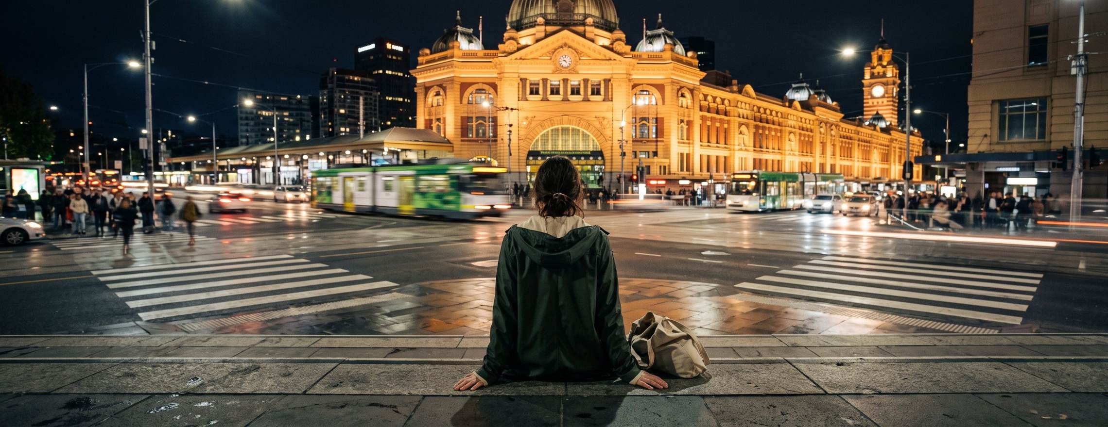 A woman seen from behind sitting still on stone steps at night, watching trams and people rush past Flinders Street Station in Melbourne