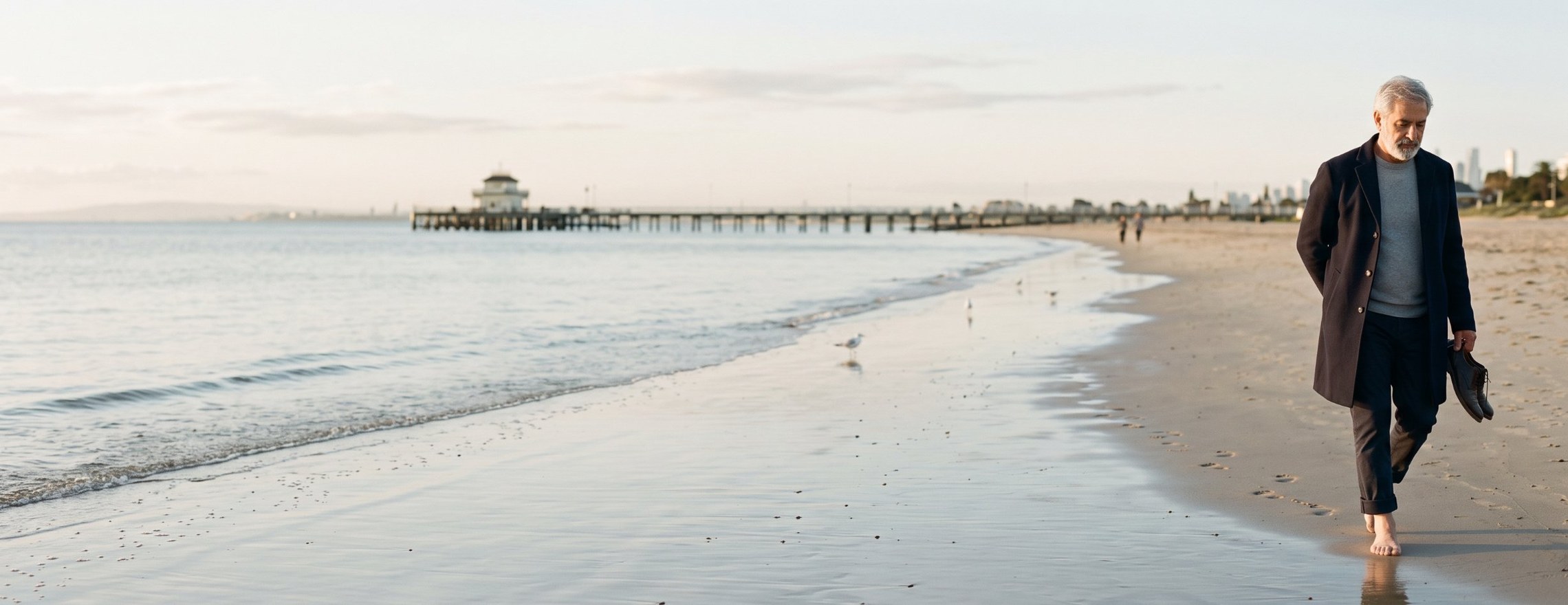An older man with grey hair walking barefoot and alone along the water at St Kilda beach in the early morning, holding his shoes, the pier in the distance