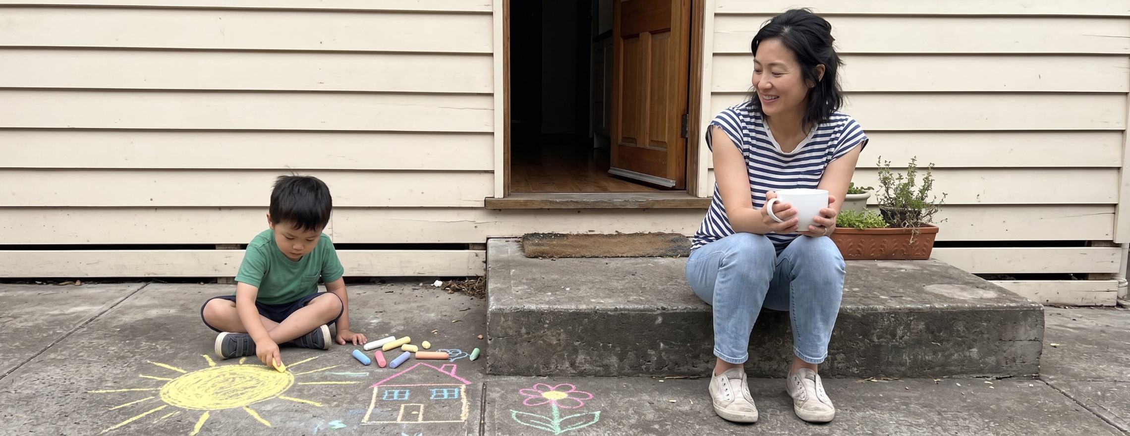Mother sitting on the back step with a mug of tea, smiling at her young son drawing with chalk
