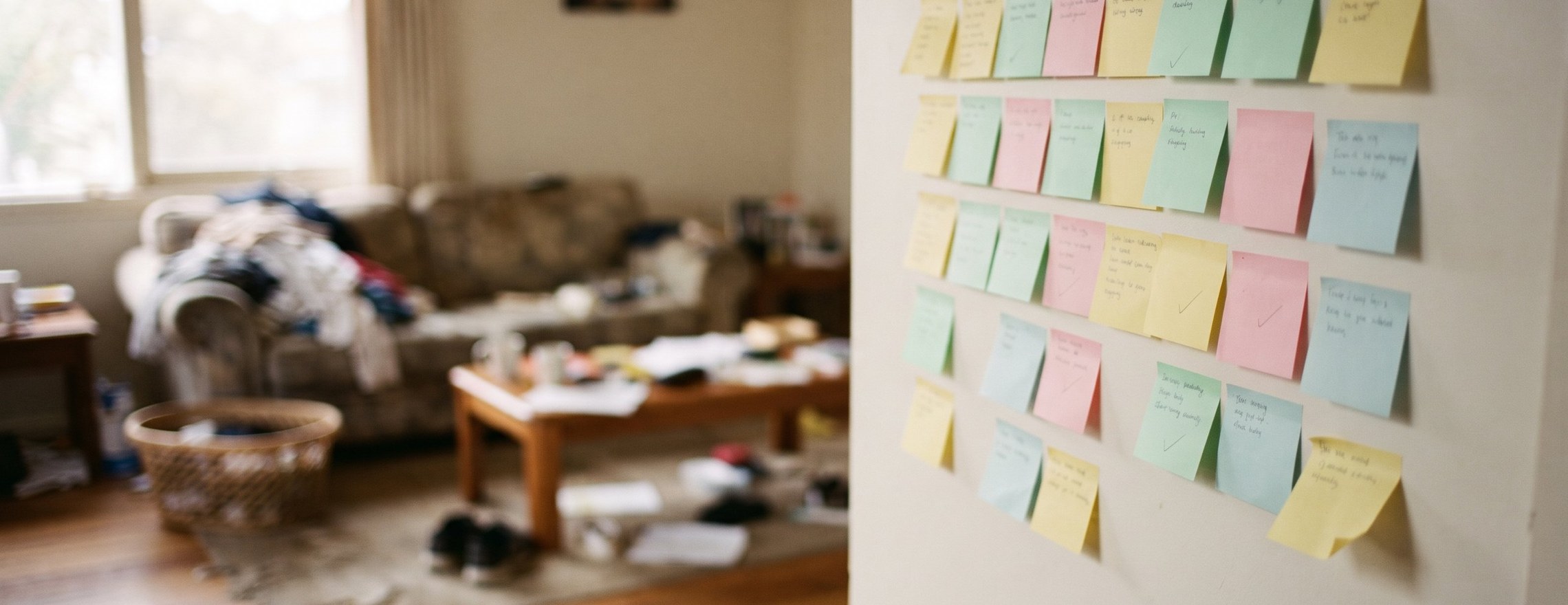 Wall covered in colourful sticky notes with a few ticked off, in front of a blurred messy living room with laundry piled on the sofa