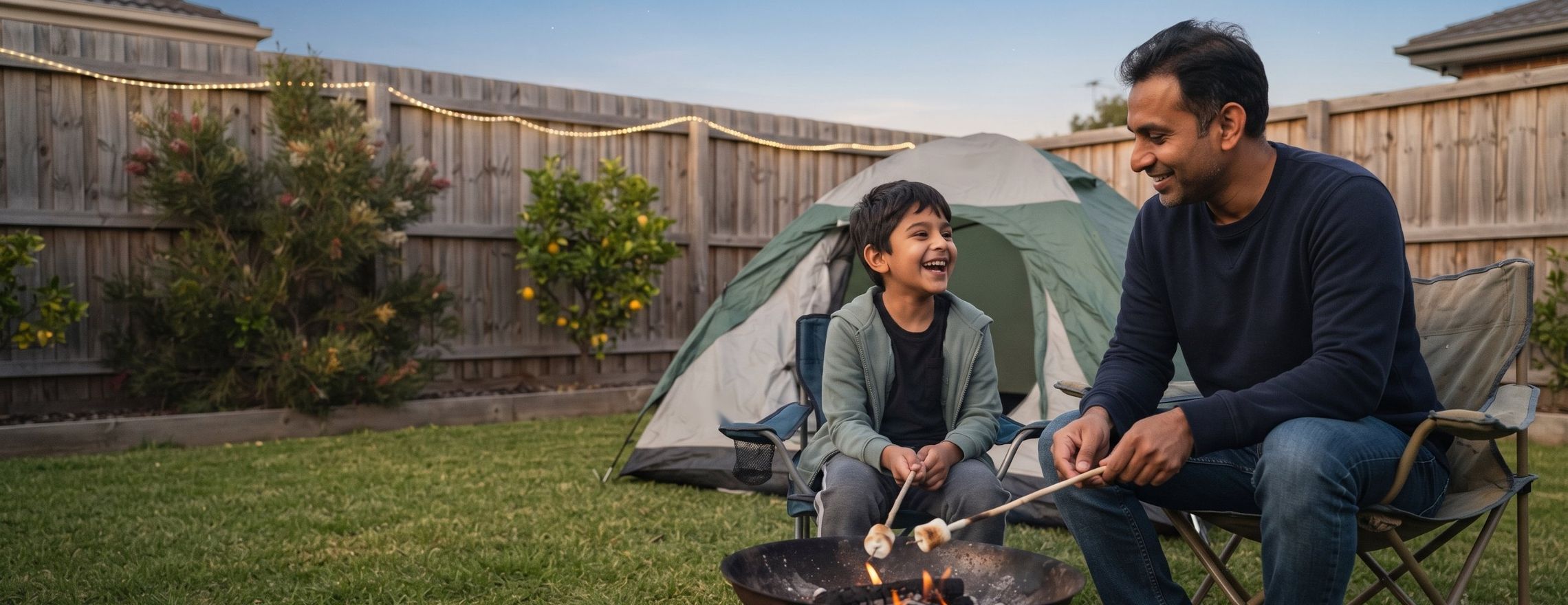 Father and son laughing as they roast marshmallows over a small fire pit beside a tent in their backyard at dusk